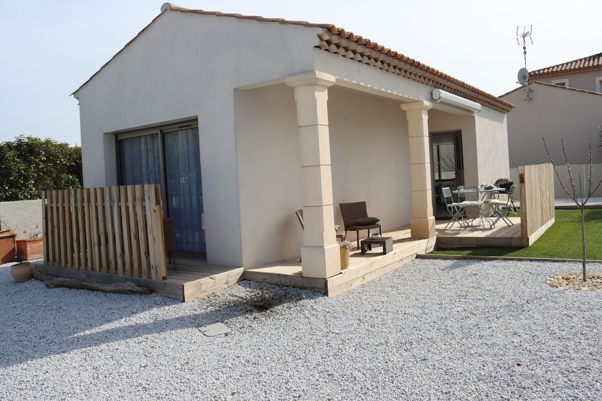A modern vacation cottage features a covered porch with two chairs positioned beside a small table. A wooden fence provides privacy, and a gravel pathway leads to the entrance. The surrounding area includes a well-maintained garden with green grass.