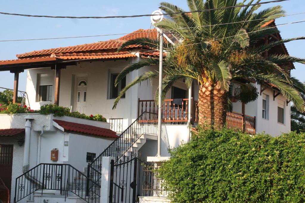 A two-story house is visible, featuring a blend of white walls and terracotta roofing. Lush palm trees and greenery surround the property, while a decorative balcony adds character. A set of stairs leads to the entrance, framed by wrought iron railings.
