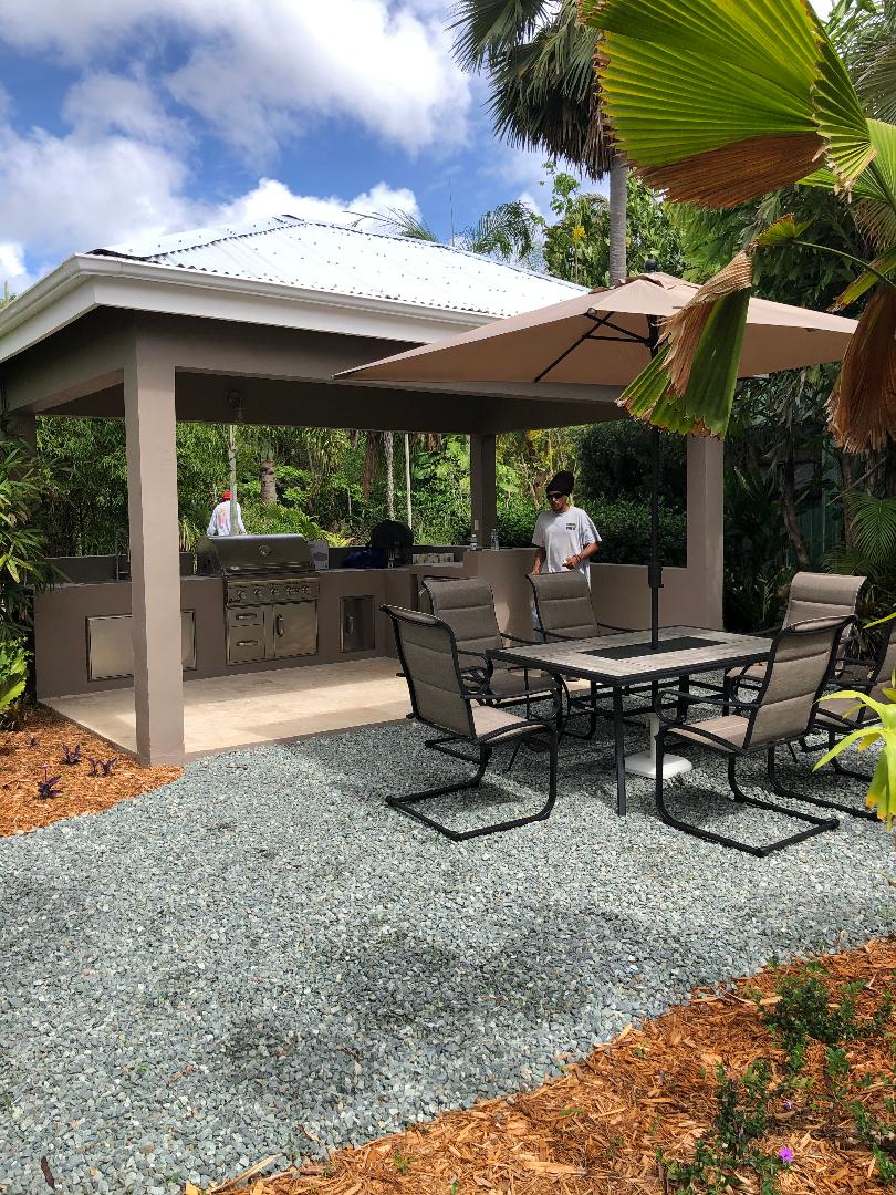 An outdoor dining area is presented under a spacious gazebo. A professional grill is positioned in the back, surrounded by a dining table with six chairs. An umbrella provides shade, while lush tropical foliage and gravel pathways enhance the tranquil setting.