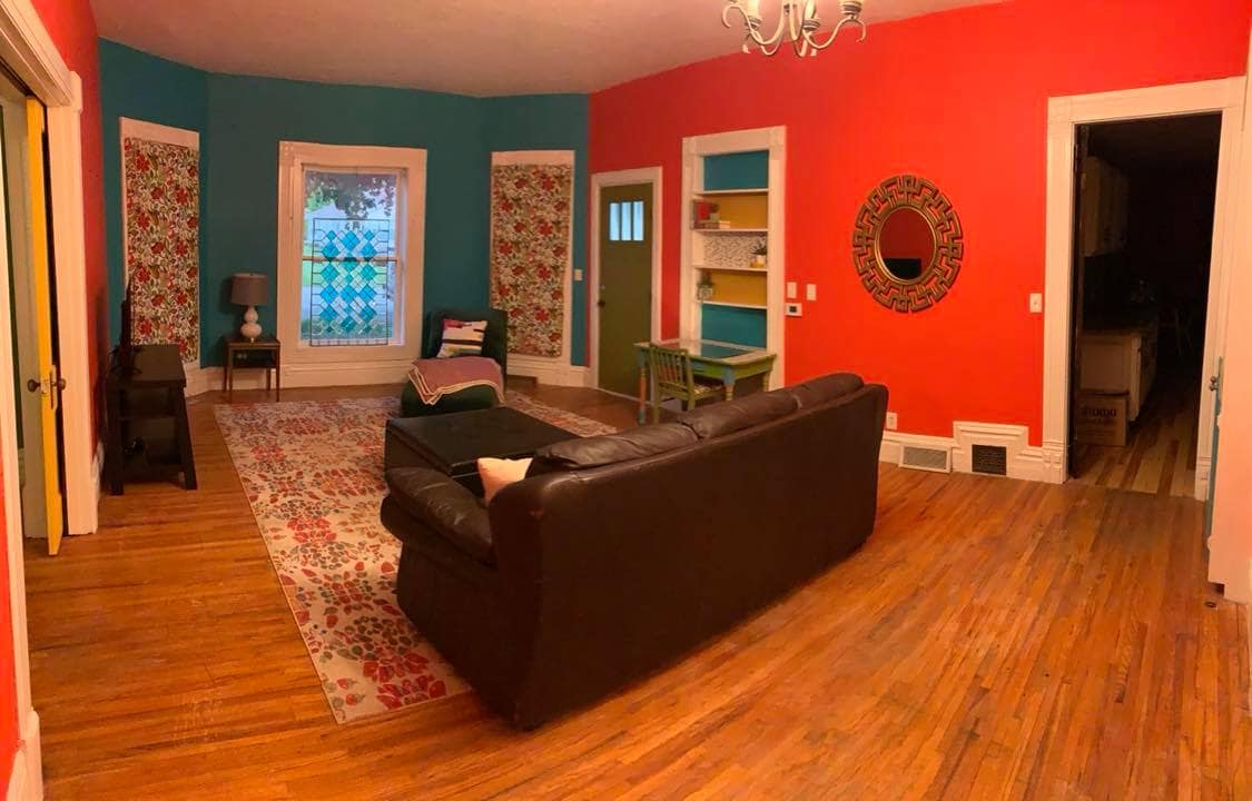 A welcoming living room features a dark leather sofa positioned on a colorful area rug. Vibrant red and teal walls are complemented by floral window treatments. Shelving with books and decor is visible, along with a round mirror reflecting light from the nearby window.