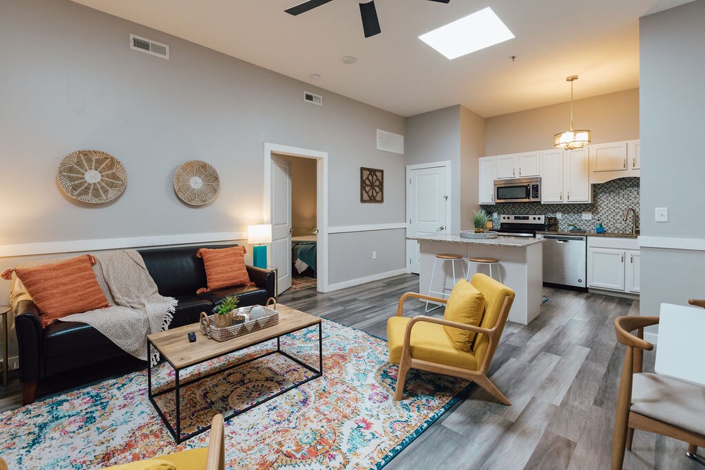A modern living area features a black couch adorned with orange and teal cushions. A patterned area rug lies beneath a wooden coffee table. A kitchen with white cabinetry and stainless-steel appliances is visible in the background, illuminated by overhead lighting.
