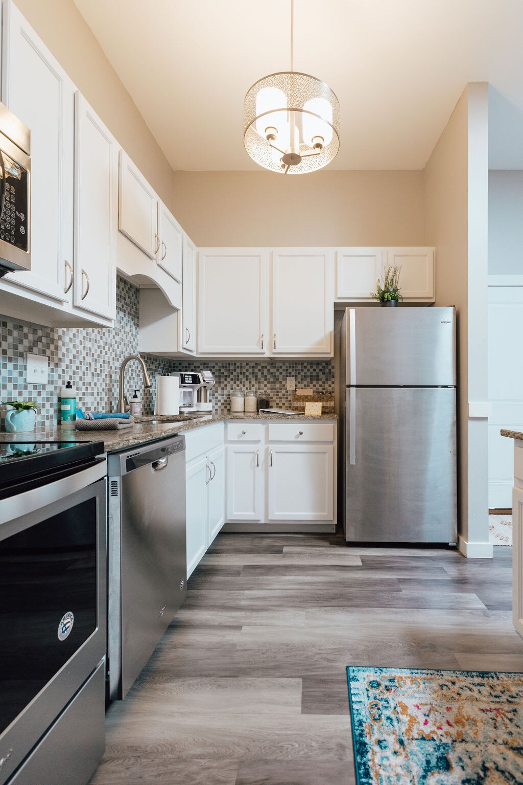 A modern kitchen features white cabinetry and stainless steel appliances, including a refrigerator and dishwasher. A mosaic tile backsplash adds visual interest, while a pendant light hangs above. The space is enhanced by a colorful area rug on the floor.