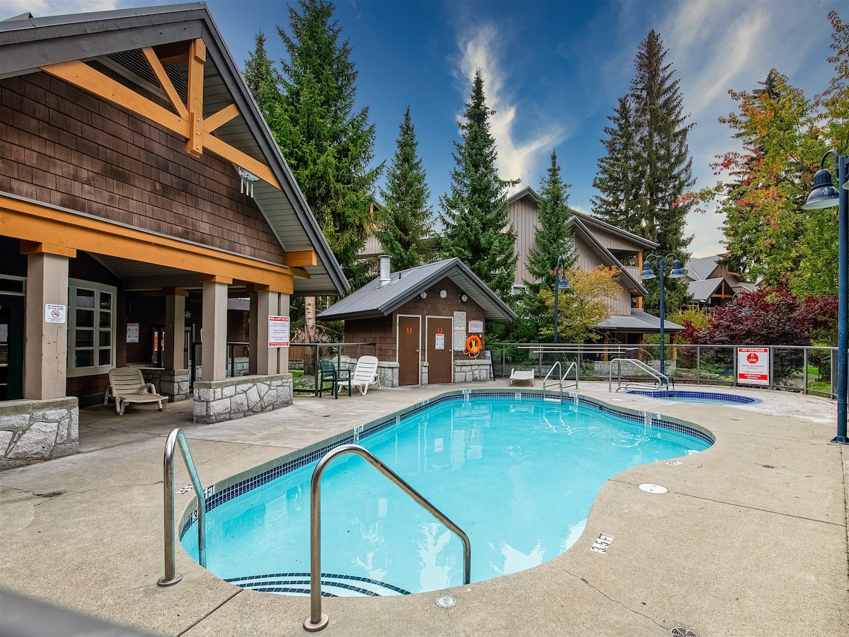 A heated outdoor pool is surrounded by landscaped greenery and modern townhomes. Comfortable lounge chairs are positioned around the pool area. Clear blue water reflects the sky, inviting relaxation in a serene setting.