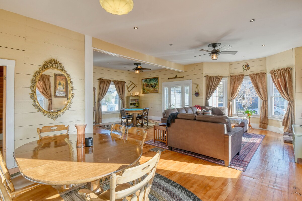 An open living area features a dining table with wooden chairs and a large cushy couch facing a window. Light enters through the draped windows, illuminating the warm wood floors. A tasteful mirror hangs on the wall, complementing the inviting decor.