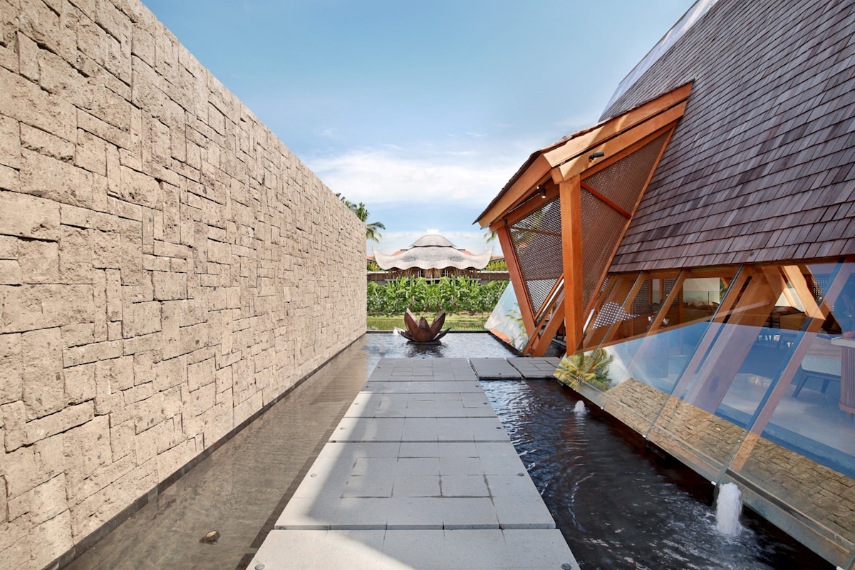 A modern architectural design is showcased, featuring a wooden structure with large glass panels and angled roofs. A stone pathway lined with water features leads to the entrance, surrounded by lush greenery and a serene atmosphere.