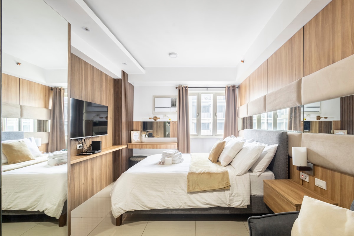 A bright and inviting bedroom features a spacious bed with neatly arranged pillows. A large mirror reflects the wood-accented walls and contemporary furniture, which includes a TV and a small workstation. Natural light filters through the window, complementing the modern design.
