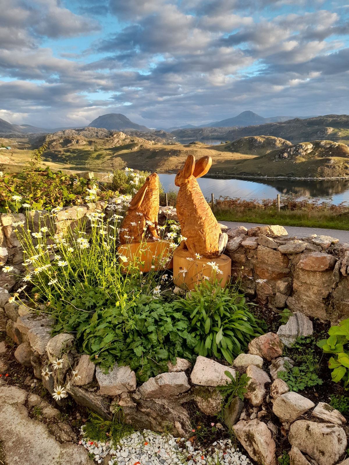 Two wooden rabbit sculptures are positioned amidst lush green plants and white flowers, surrounded by a circular stone border. A scenic landscape of hills and a shimmering lake is visible in the background, enhanced by dynamic cloud patterns in the sky.