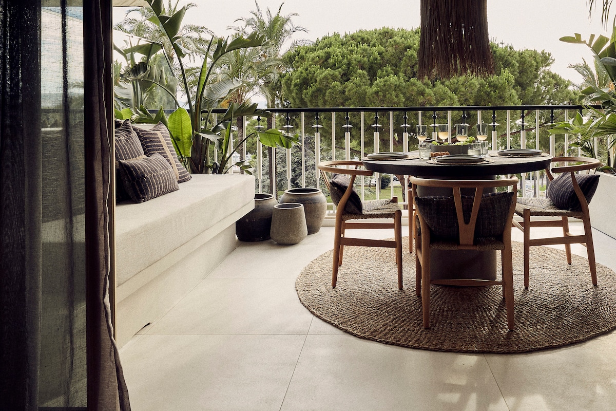 A spacious terrace is adorned with a round dining table surrounded by four wooden chairs. A lounge area features a long, cushioned sofa along the railing, complemented by decorative pillows. Lush greenery and palm trees create a serene backdrop, enhancing the outdoor setting.