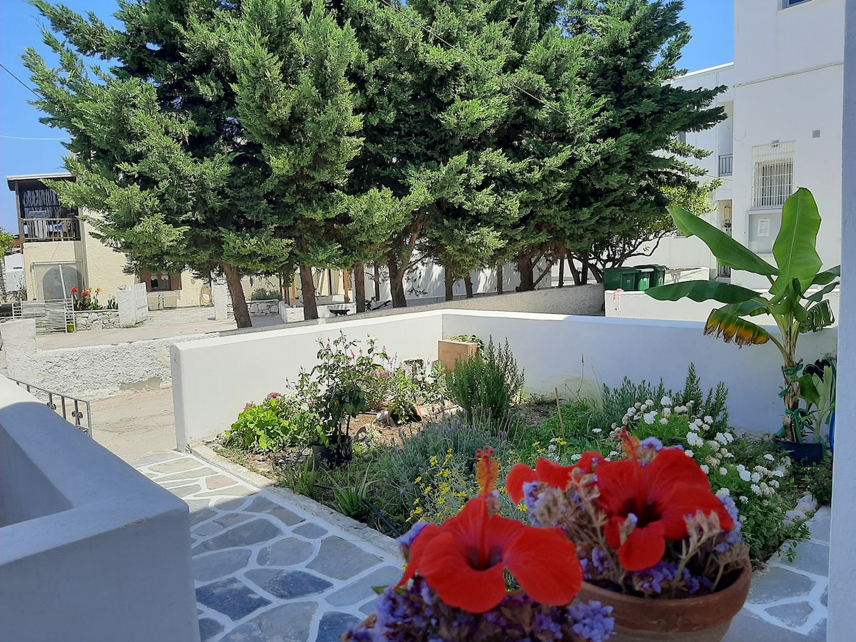 A garden area is shown featuring colorful flowers in the foreground, including red blooms. Lush greenery is visible in the background with tall trees providing shade. The space is surrounded by a low white wall and stone flooring, suggesting a peaceful outdoor environment.