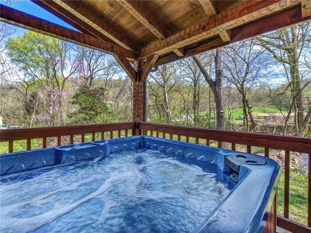 A large hot tub with bubbling water is set under a wooden roof on a covered deck. Surrounding trees provide a natural backdrop, and glimpses of the landscape can be seen through the railing, creating a serene atmosphere for relaxation.