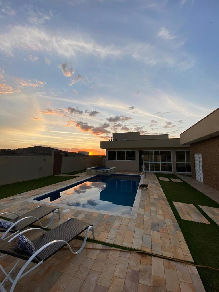 The image captures a modern home with a spacious pool area under a colorful sunset. Lounge chairs are positioned beside the pool, which reflects the sky's hues. The home features large windows, allowing for a view of the outdoors, enhancing the tranquil atmosphere.
