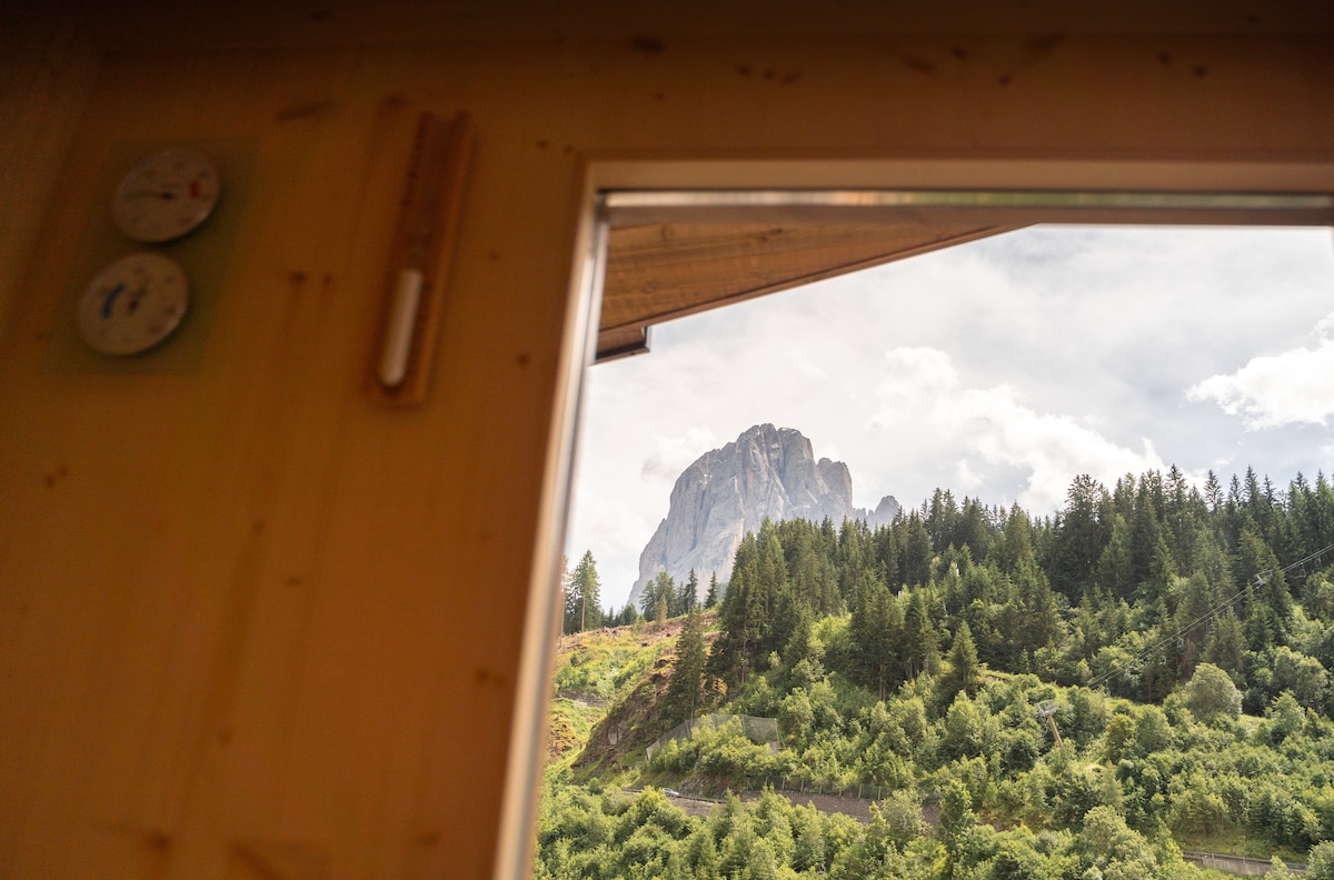 A view through a window highlights the surrounding natural landscape, featuring lush greenery and towering mountains. The scene is framed by wooden panels, suggesting a cozy interior. Weather instruments are visible on the wall near the window, adding a functional touch to the setting.