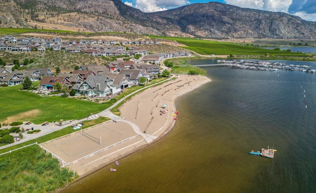 An aerial view captures the sandy beach and calm waters of Osoyoos Lake, bordered by a grassy area featuring volleyball courts. The beach is lined with colorful umbrellas and kayaks, while the backdrop showcases the community's elegant homes and surrounding hills.