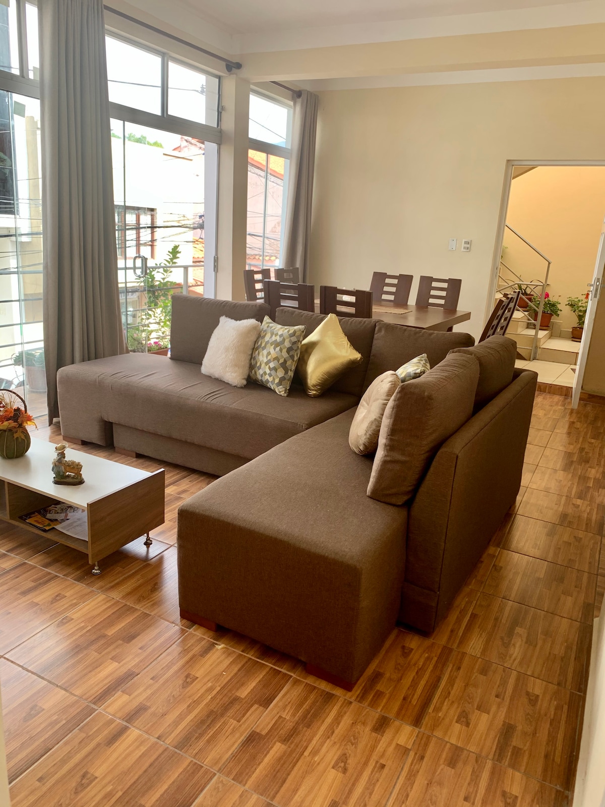 A cozy living area features a large sectional sofa in neutral tones, adorned with decorative pillows. A small coffee table is situated in front, while a dining table with multiple chairs is visible in the background. Natural light floods in through the large windows.