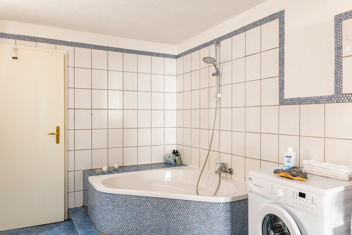 The spacious bathroom features a corner bathtub with a shower fixture mounted on the wall. A washing machine is positioned nearby, and the area is tiled with light blue and white squares. Essentials are neatly arranged on shelves and surfaces.