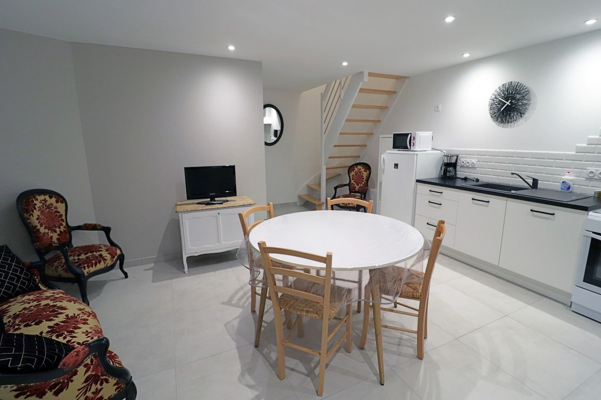 An open-plan kitchen and living area is displayed, featuring a round dining table surrounded by wooden chairs. A comfortable armchair is positioned nearby, while a television sits on a small cabinet. The kitchen is equipped with essential appliances against a white wall, and a staircase is visible in the background.