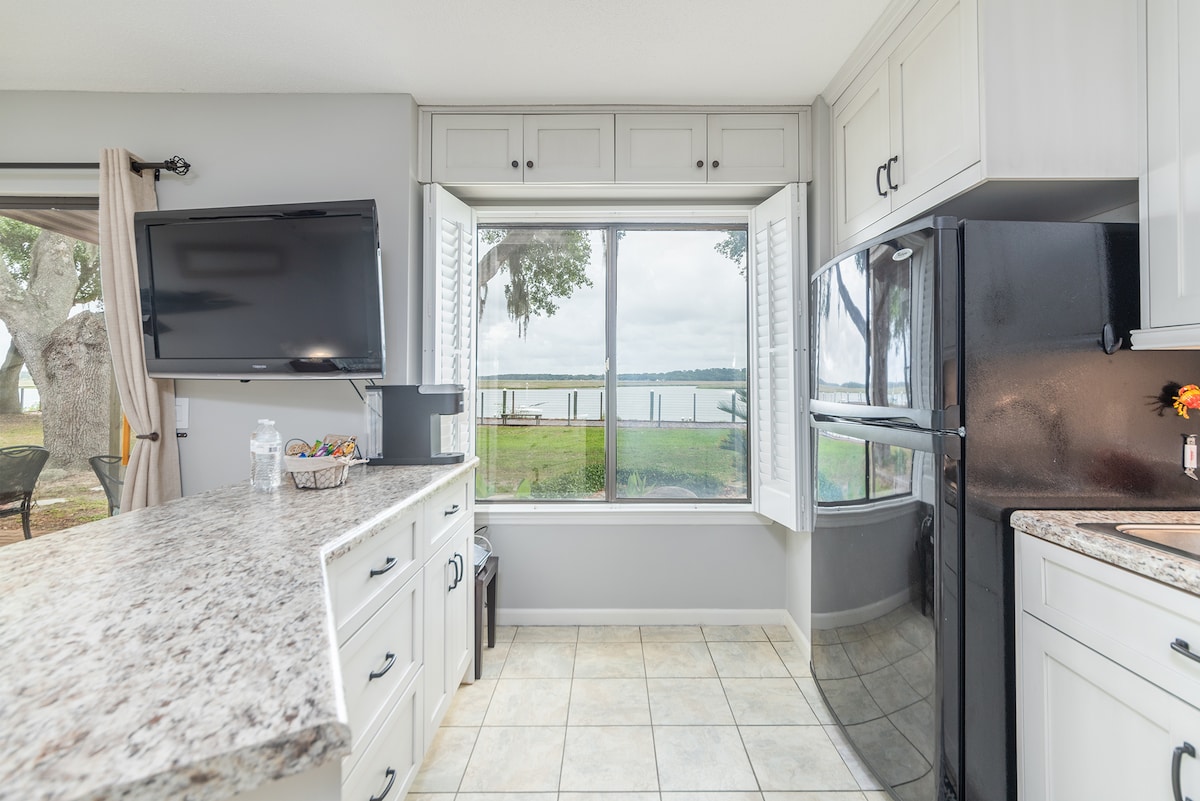 A light-filled kitchen features a large granite countertop with ample workspace and modern appliances. Windows wrap around the space, offering views of the water, while a television is mounted for entertainment. A bowl of fresh fruit adds a welcoming touch, enhancing the functional kitchen layout.