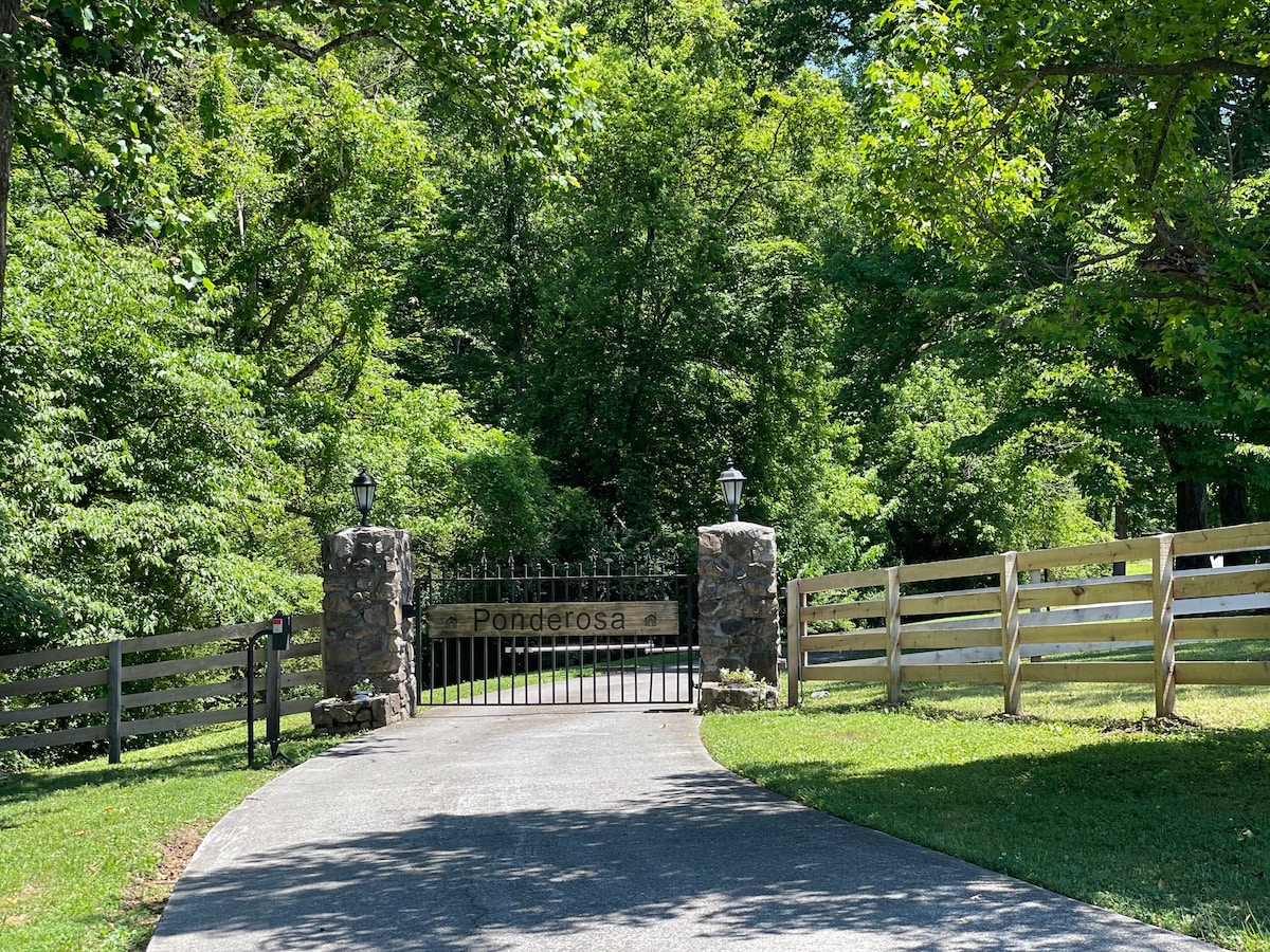 A scenic entryway features a stone gate with a sign reading 'Ponderosa,' framed by lush greenery and trees lining the entrance. A paved driveway leads into the tranquil retreat, bordered by a wooden fence and well-maintained grass.