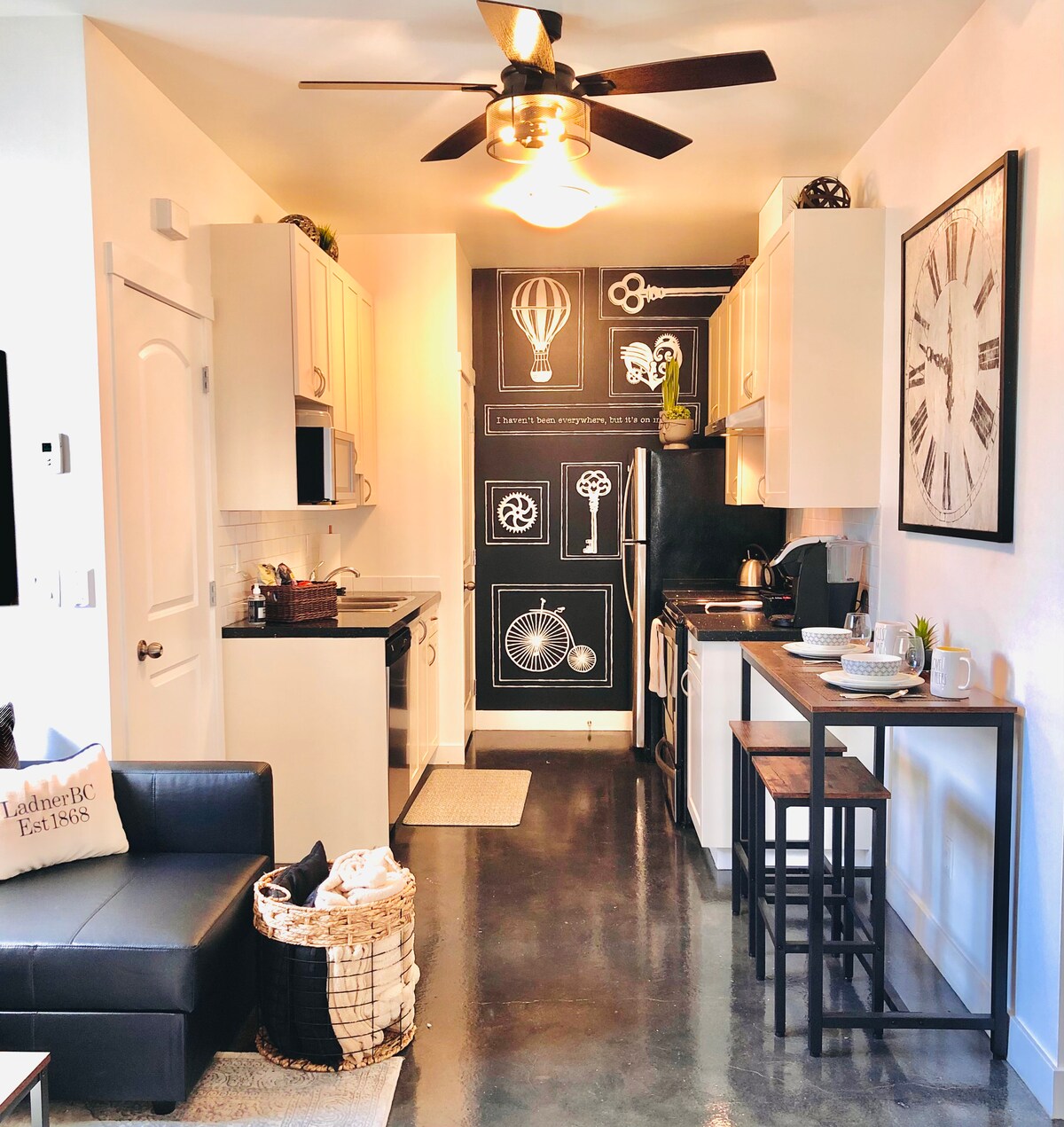 An open kitchen area features modern appliances including a stove and refrigerator, complemented by sleek cabinetry. A black accent wall displays stylish graphic designs. A cozy couch and small dining table are visible, enhancing the space's functionality. Warm light emanates from the ceiling fan.
