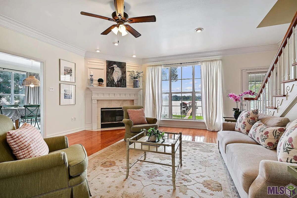 A bright living area features a mix of seating, including a green armchair and a floral-patterned sofa. A coffee table sits at the center, with a fireplace and large windows providing views of the lake, framed by sheer drapes that allow natural light to illuminate the space.