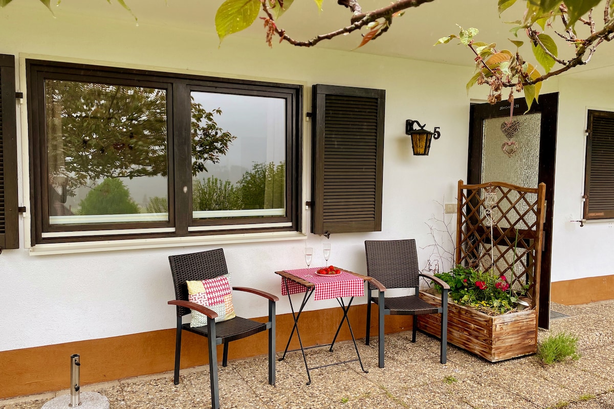 A cozy outdoor seating area is highlighted by two chairs positioned around a small table. A decorative flower box adds a touch of greenery, while a window with shutters above provides a view of the surroundings. The entrance is framed by a small trellis, enriching the scene.