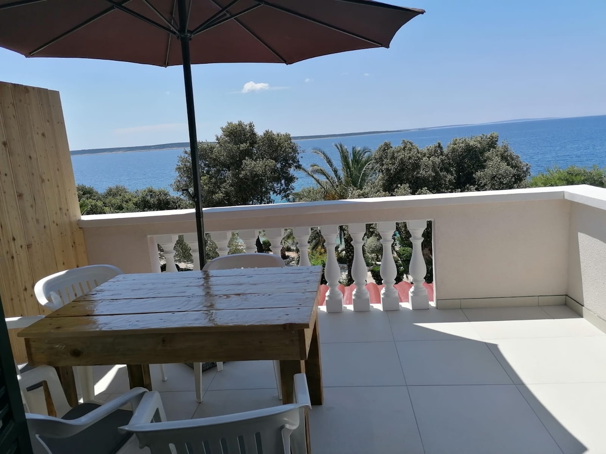 An outdoor terrace features a wooden table surrounded by white plastic chairs. A large umbrella provides shade, while the view overlooks the blue sea and lush greenery beyond the railing.