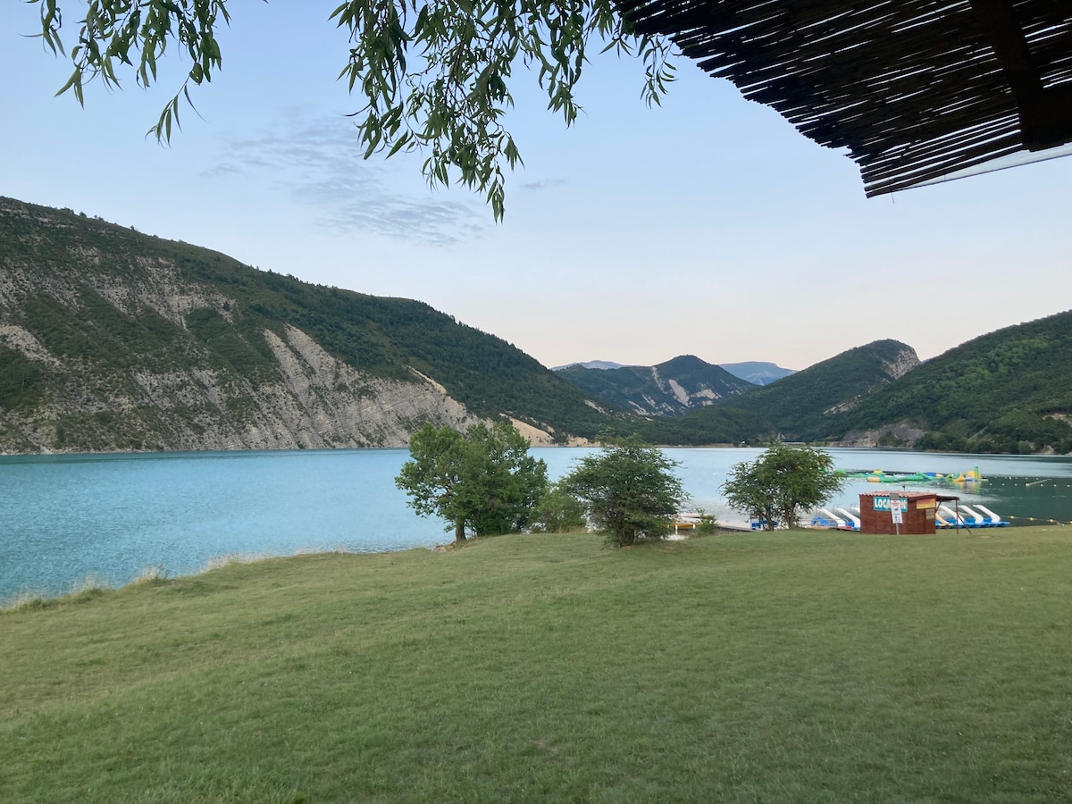 A serene lakeside view captures the turquoise water nestled between green hills. A grassy area leads to the water's edge, where various water activities and inflatable structures are visible. The scene is framed by trees, enhancing the natural beauty of the landscape.