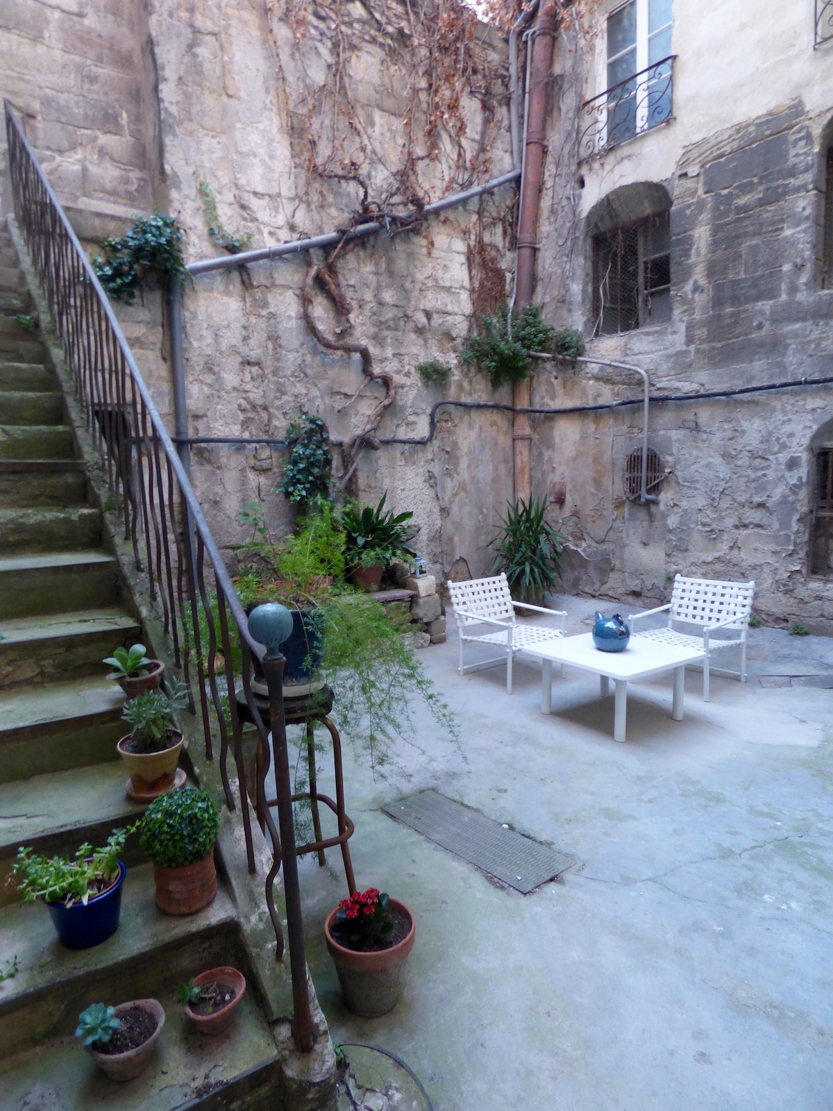 A shared courtyard features stone walls adorned with climbing plants and small potted flowers. Several chairs surround a white table, creating a seating area. A staircase leads to an upper level, framed by a wrought iron railing, enhancing the serene outdoor space.