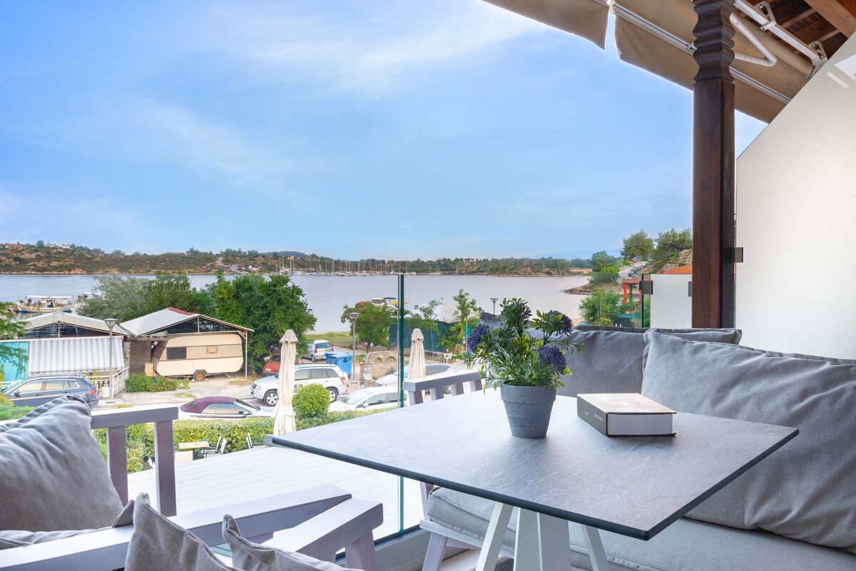 A balcony seating area features plush gray cushions on white furniture, offering views of the water and surrounding marina. A small potted plant adorns the table, while the landscape showcases boats docked along the shore, enhancing the serene atmosphere.