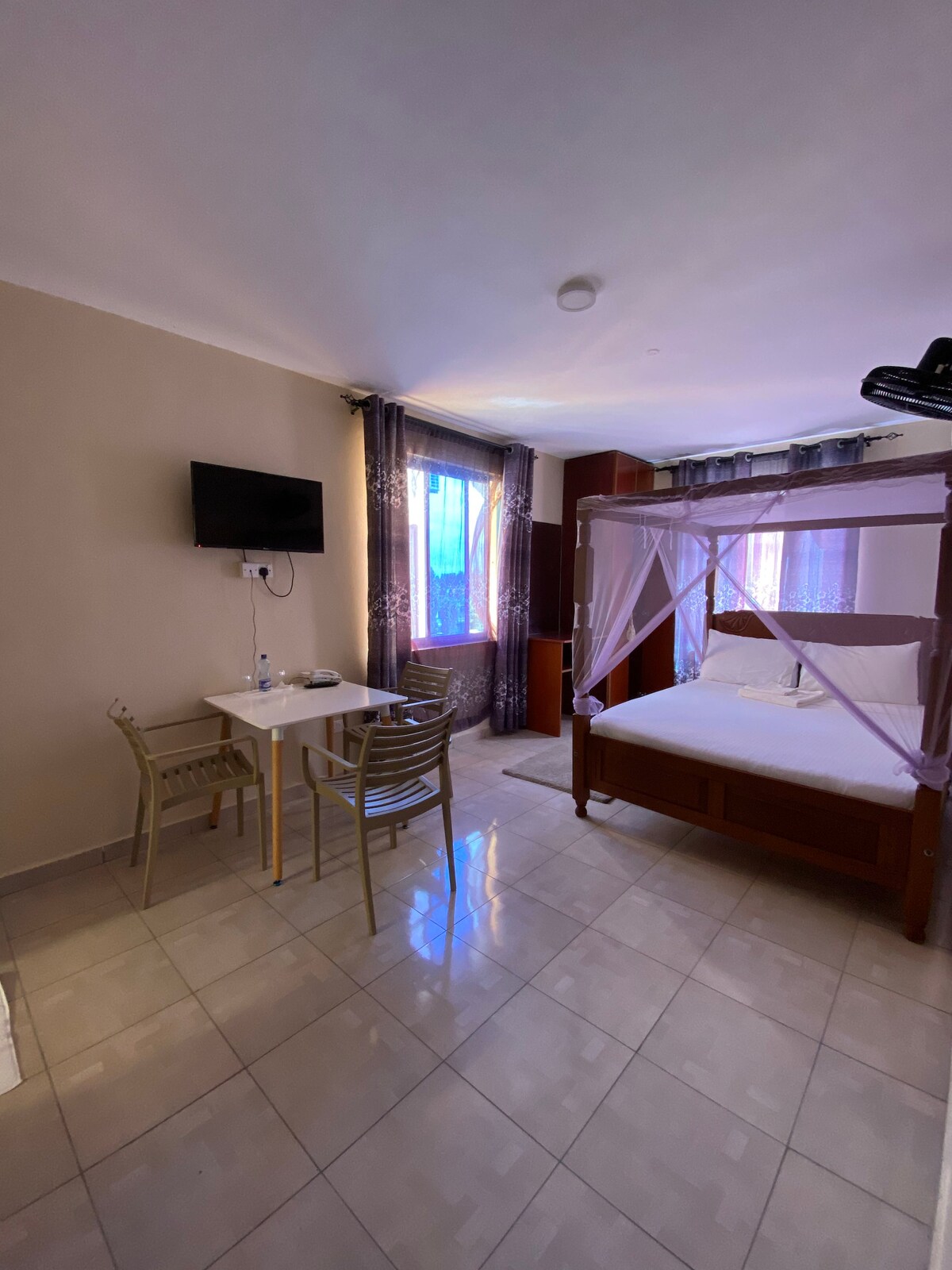 A bright room features a spacious bed with a white canopy, accompanied by a small dining table with two chairs. Natural light enters through two windows, with sheer curtains adding a soft touch. A wall-mounted television is visible, enhancing the room's functionality.