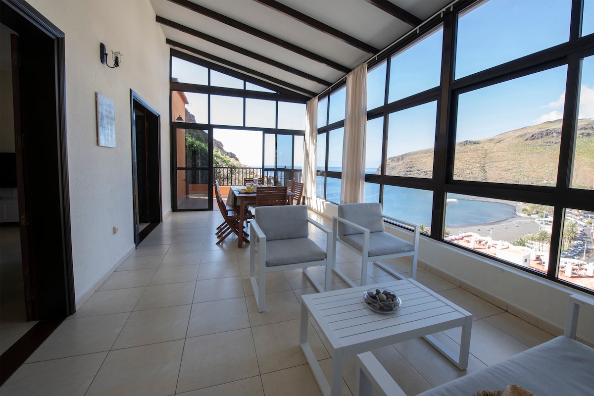 A bright, glass-enclosed terrace features a seating area with white furniture, offering panoramic views of the bay and mountains. Natural light floods the space through large windows, enhancing the inviting atmosphere for relaxation or dining.
