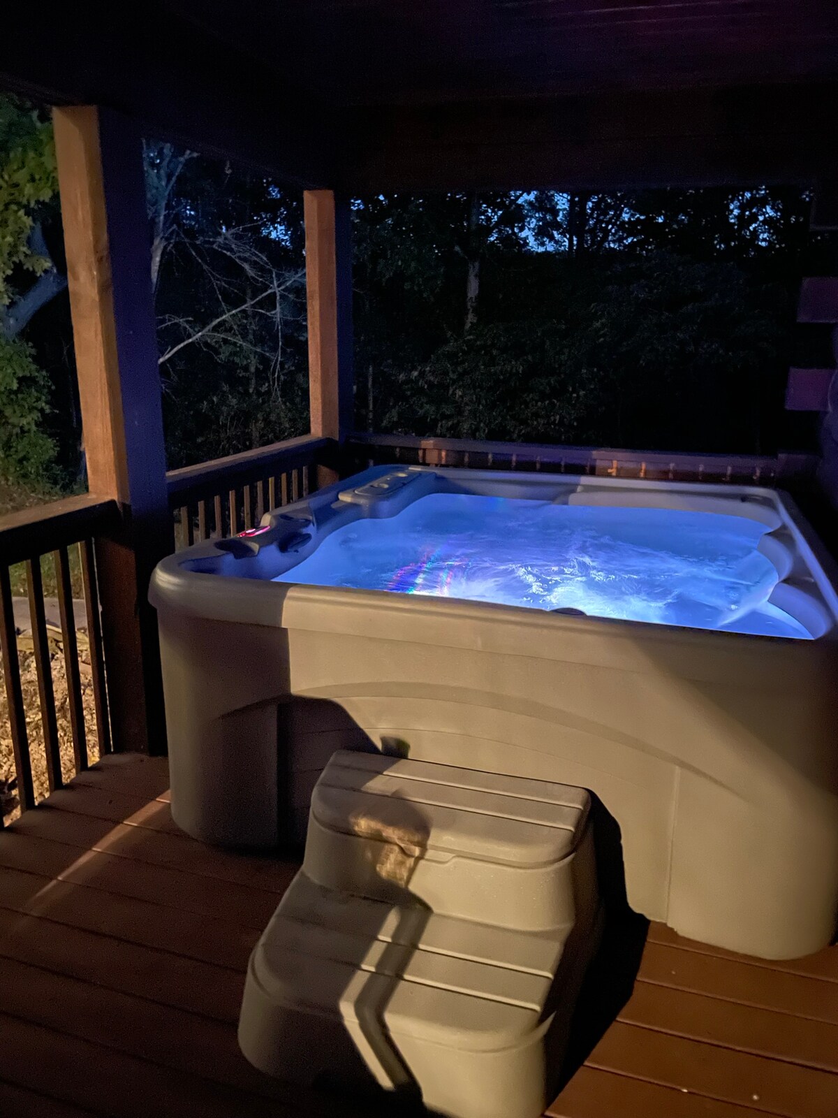 A hot tub is positioned on a wooden deck, softly illuminated with blue light. Steps lead up to the tub, surrounded by natural greenery, creating a serene outdoor space for relaxation.