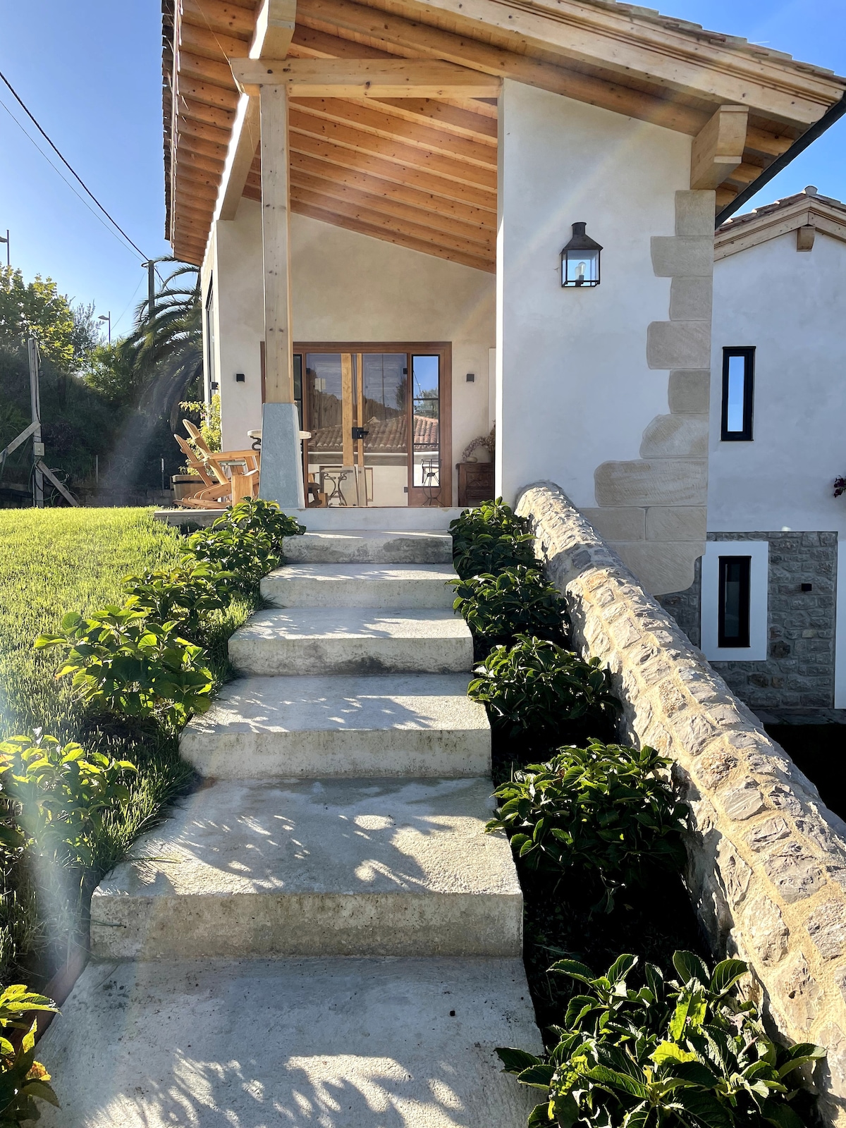A welcoming entrance features a stone pathway leading to the front door, framed by elegant greenery and natural landscaping. The structure showcases a sloped roof with wooden beams and a lantern, inviting guests to enter the home.