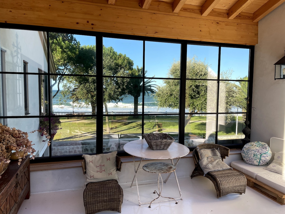 A sunlit seating area features large windows showcasing a view of the sea and surrounding greenery. A round table is accompanied by wicker chairs, inviting relaxation. The ceiling is adorned with wooden beams, enhancing the space's character.