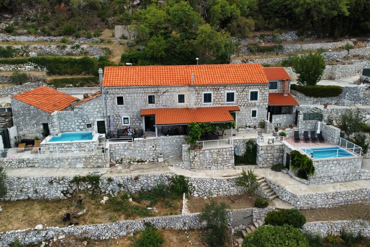 An aerial view showcases a traditional countryside stone house with a terracotta roof. Private pools are visible next to a spacious patio area, surrounded by stone walls and lush greenery, creating an inviting atmosphere. The landscape features cultivated fields and trees in the background.
