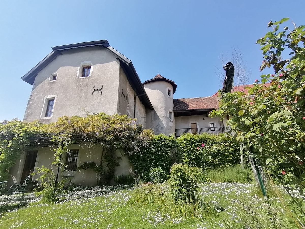 A historic château is surrounded by lush greenery and blooming flowers, highlighting its rustic charm. The building features a stone exterior with a unique turret and a balcony. A well-maintained garden extends at the forefront, offering a serene outdoor space.