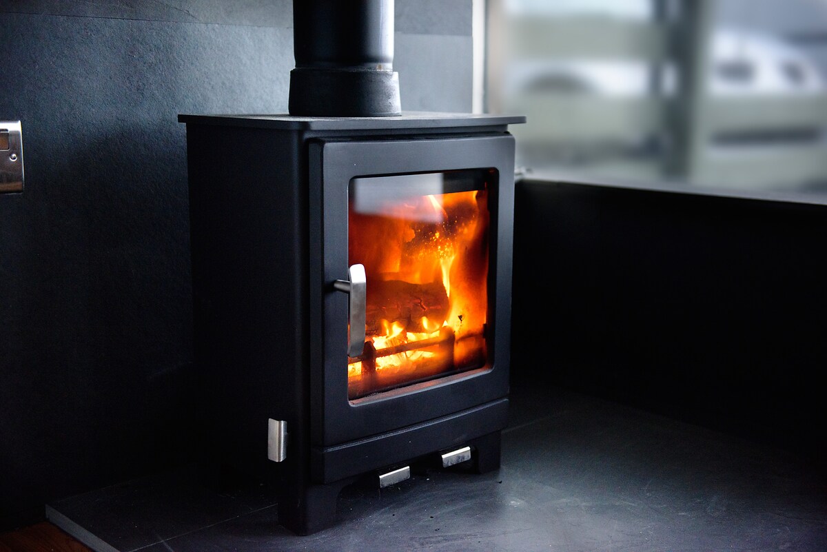 A log woodburner occupies a corner of the room, radiating warmth with a visible flame through the glass door. The stove's sleek black exterior contrasts with the surrounding surfaces, enhancing a cozy ambiance in the space.