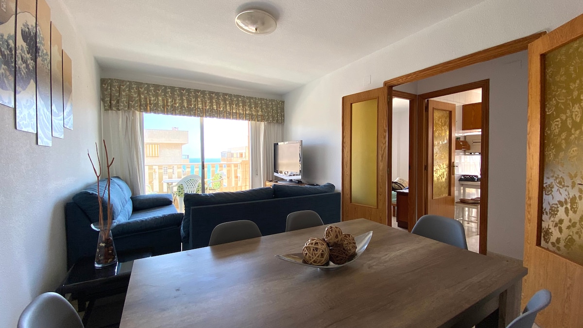 A spacious living area is presented with a large sectional sofa and a wooden dining table set for meals. Natural light filters through sheer curtains, illuminating the room. A television is positioned near the window, which offers views of the surroundings.