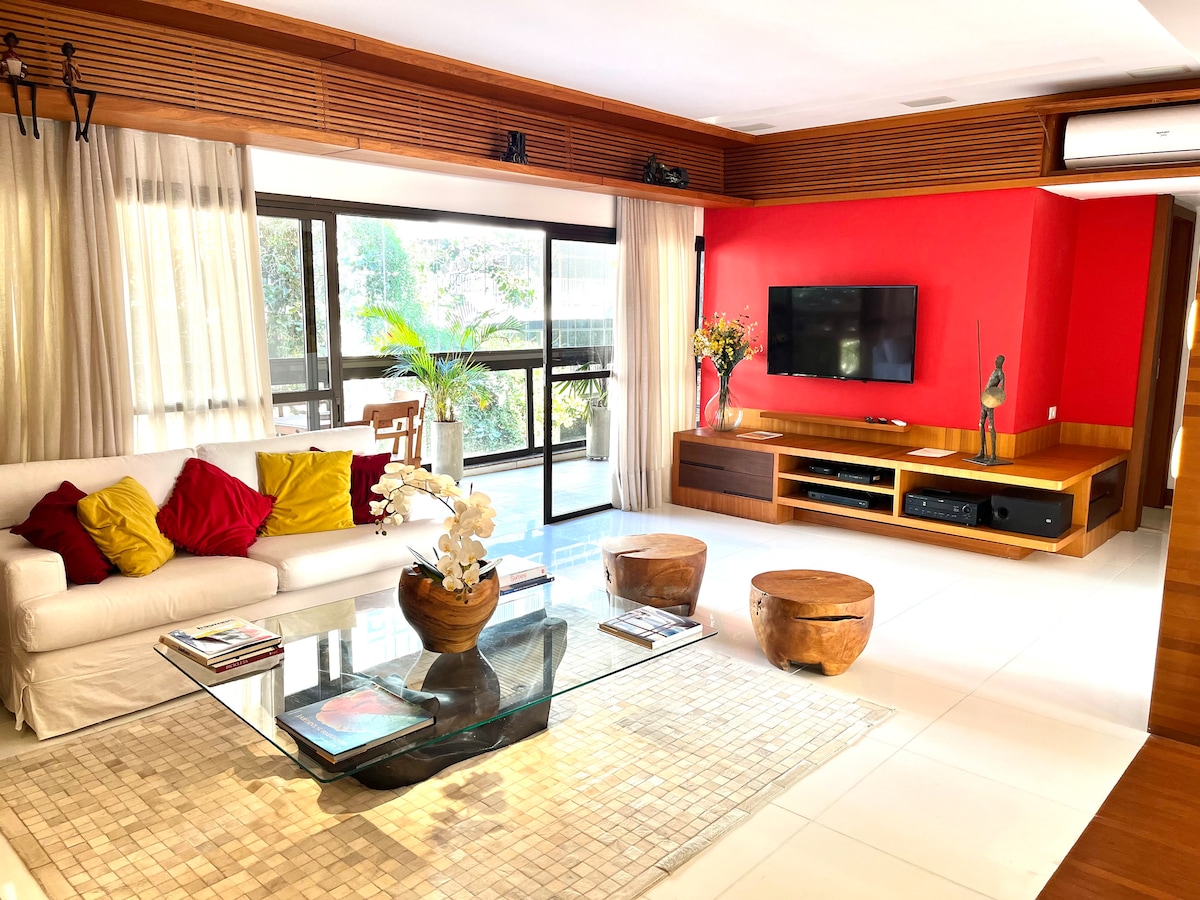 A spacious living area features a white sofa adorned with colorful cushions. A glass coffee table sits on a textured area rug, complemented by wooden accent tables. Large windows invite natural light, while a vibrant red accent wall enhances the modern design.