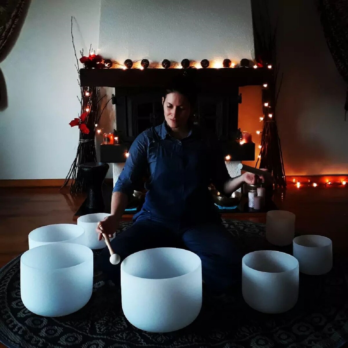 A calming indoor space is showcased, featuring several crystal singing bowls arranged on a patterned rug. Soft lighting creates a serene ambiance, with decorative elements enhancing the tranquil setting. A person is seated at the center, holding a mallet, contributing to the peaceful atmosphere.