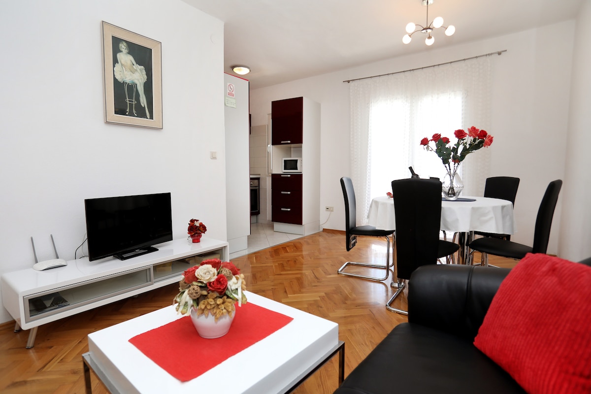 A modern living area features a sleek television on a low-profile white media unit. A black leather sofa with a striking red cushion offers seating, while a round dining table is surrounded by black chairs. A vibrant floral centerpiece adds a touch of color to the space.