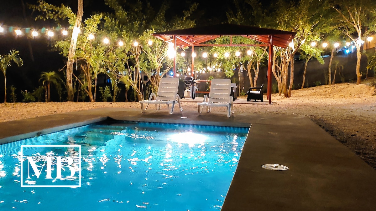 A private pool glimmers under the night sky, surrounded by a spacious patio area. Two lounge chairs are positioned beside the pool, while a covered BBQ area, illuminated by string lights, creates a welcoming setting for relaxation or dining.
