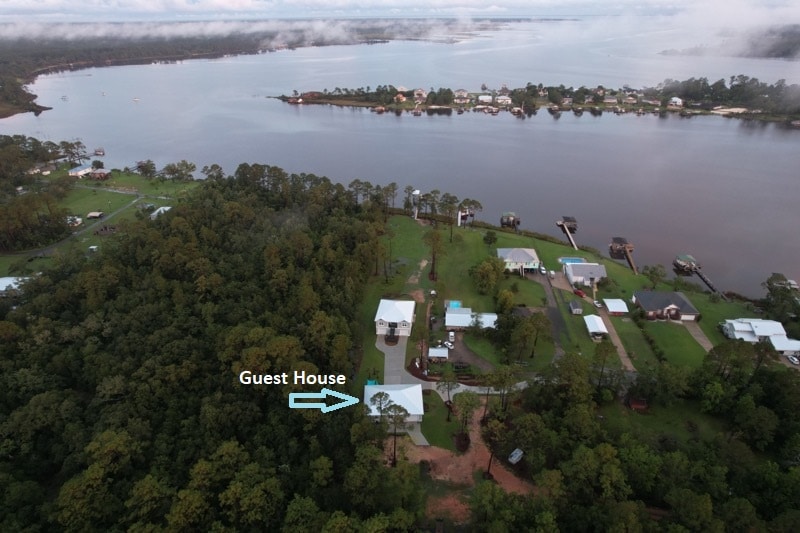 An aerial view captures a spacious waterfront estate with the guest house marked for easy identification. Lush greenery surrounds the property, providing a natural buffer. The calm waters of the bay reflect the surrounding homes and trees, illustrating the tranquil environment.