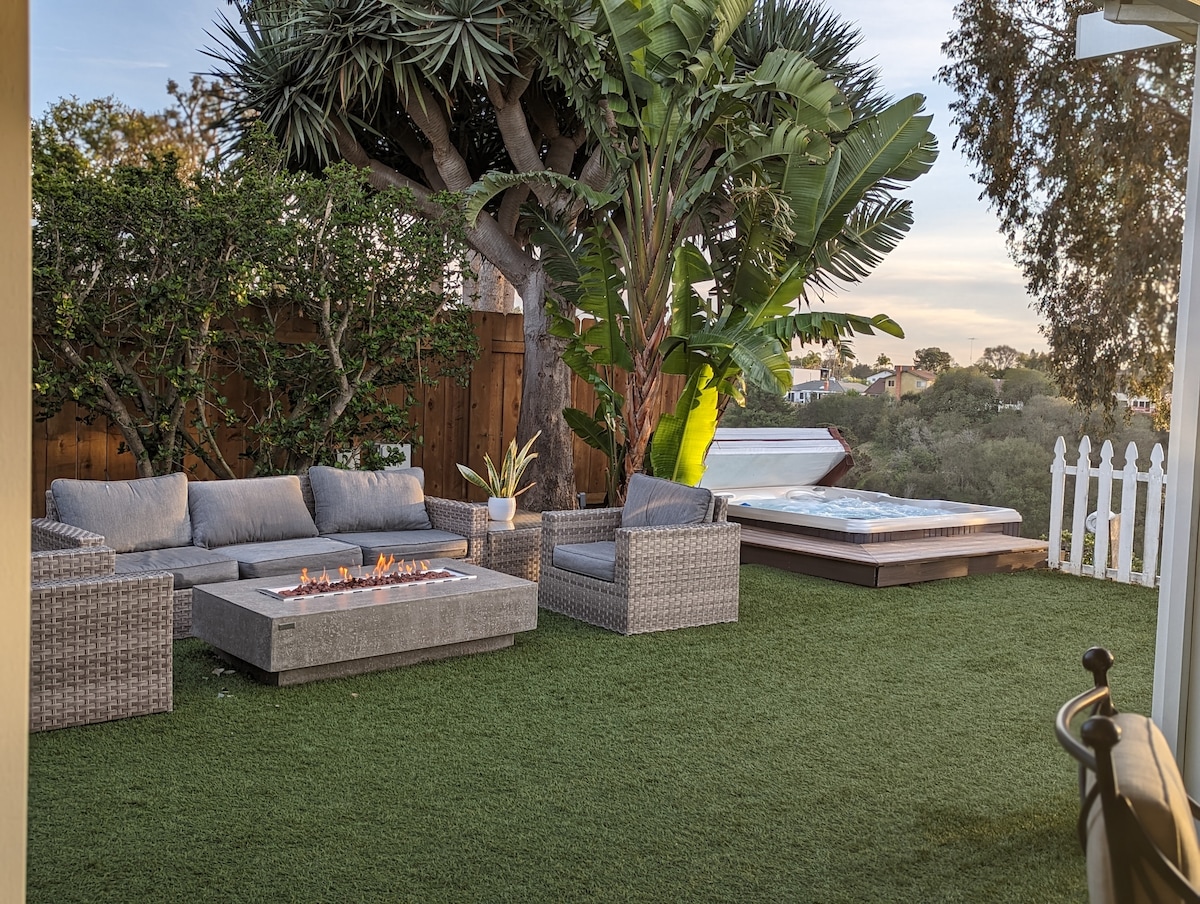 A spacious backyard features a comfortable outdoor sofa and two armchairs arranged around a fire pit. A hot tub is positioned nearby, surrounded by lush greenery and palm trees. The area is designed with artificial grass, creating a welcoming outdoor space.