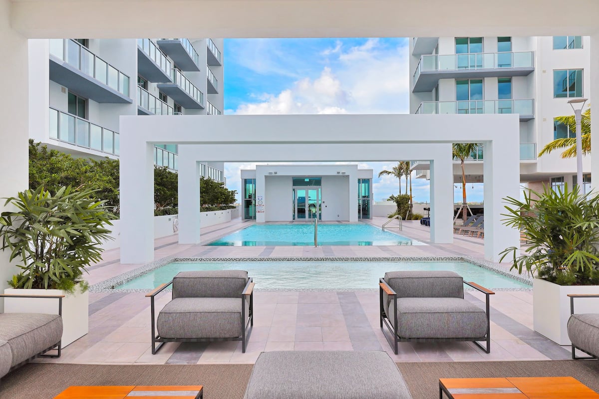 An inviting outdoor pool area features a modern design with lounge seating and palm trees. The pool is surrounded by stylish architecture, providing a serene space for relaxation. Large glass doors offer a view of the building's interior, enhancing the bright and open atmosphere.