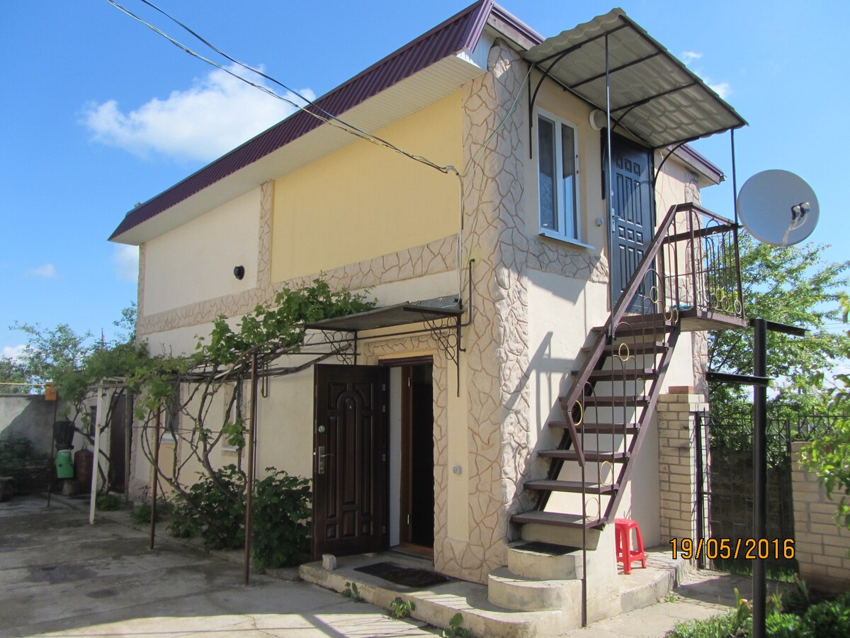A two-story guest house is depicted, featuring a stone facade and a metal staircase leading to the upper level. A small covered balcony is visible above the entrance, and outdoor seating is arranged in a paved area surrounded by greenery.