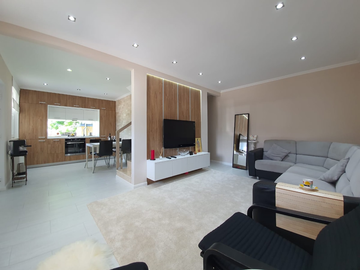 A spacious living area features a light-colored carpet and elegant wooden accents. A modern television is mounted on the wall, and seating arrangements include a gray sectional sofa and a black armchair. A dining area with four chairs is visible in the background, leading to the kitchen.