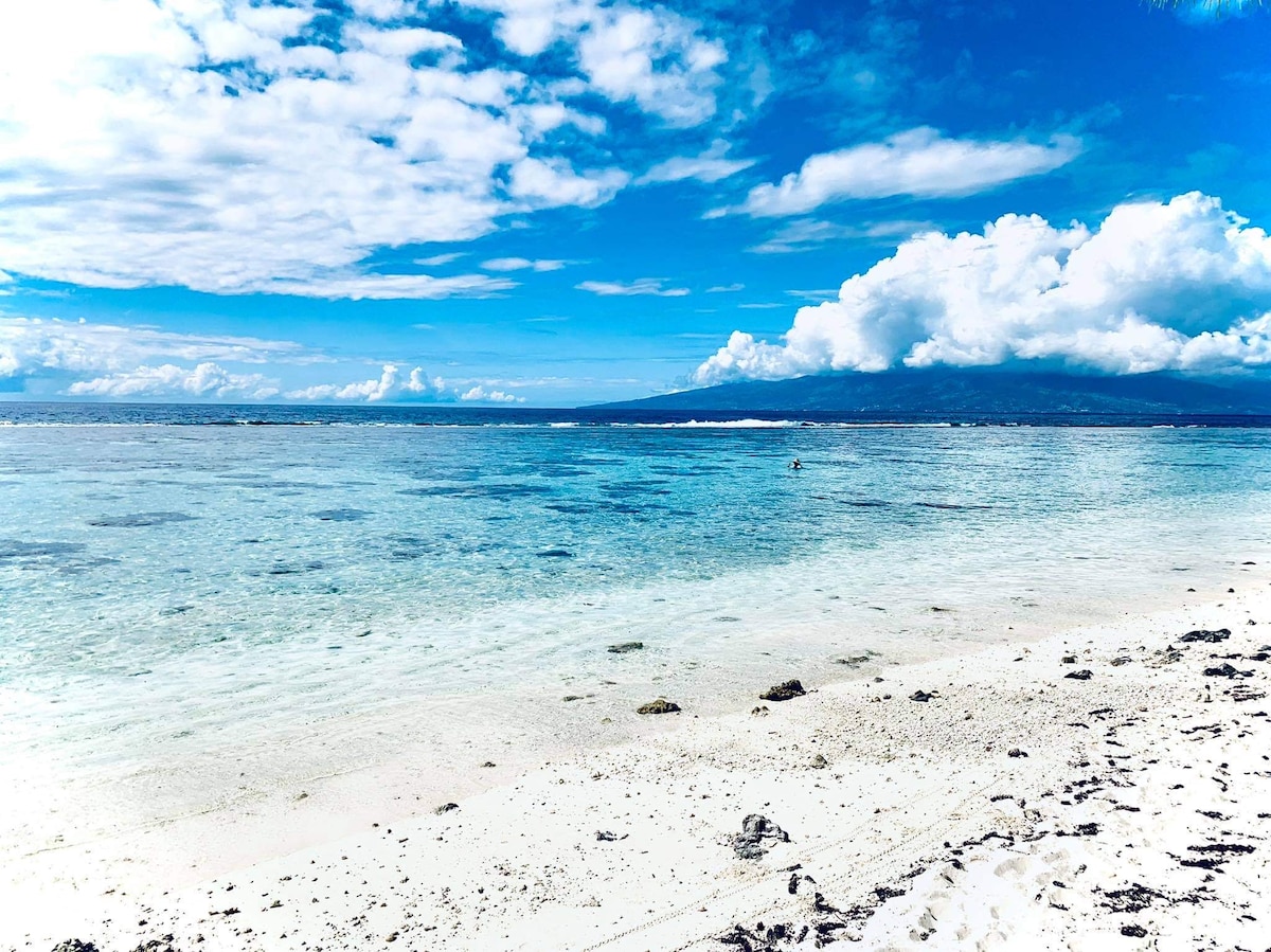 A serene beach scene reveals a clear, shallow lagoon leading to a distant island, framed by a vibrant blue sky dotted with white clouds. The sandy shoreline shows gentle waves lapping at the water's edge, enhancing the tranquil atmosphere of the environment.