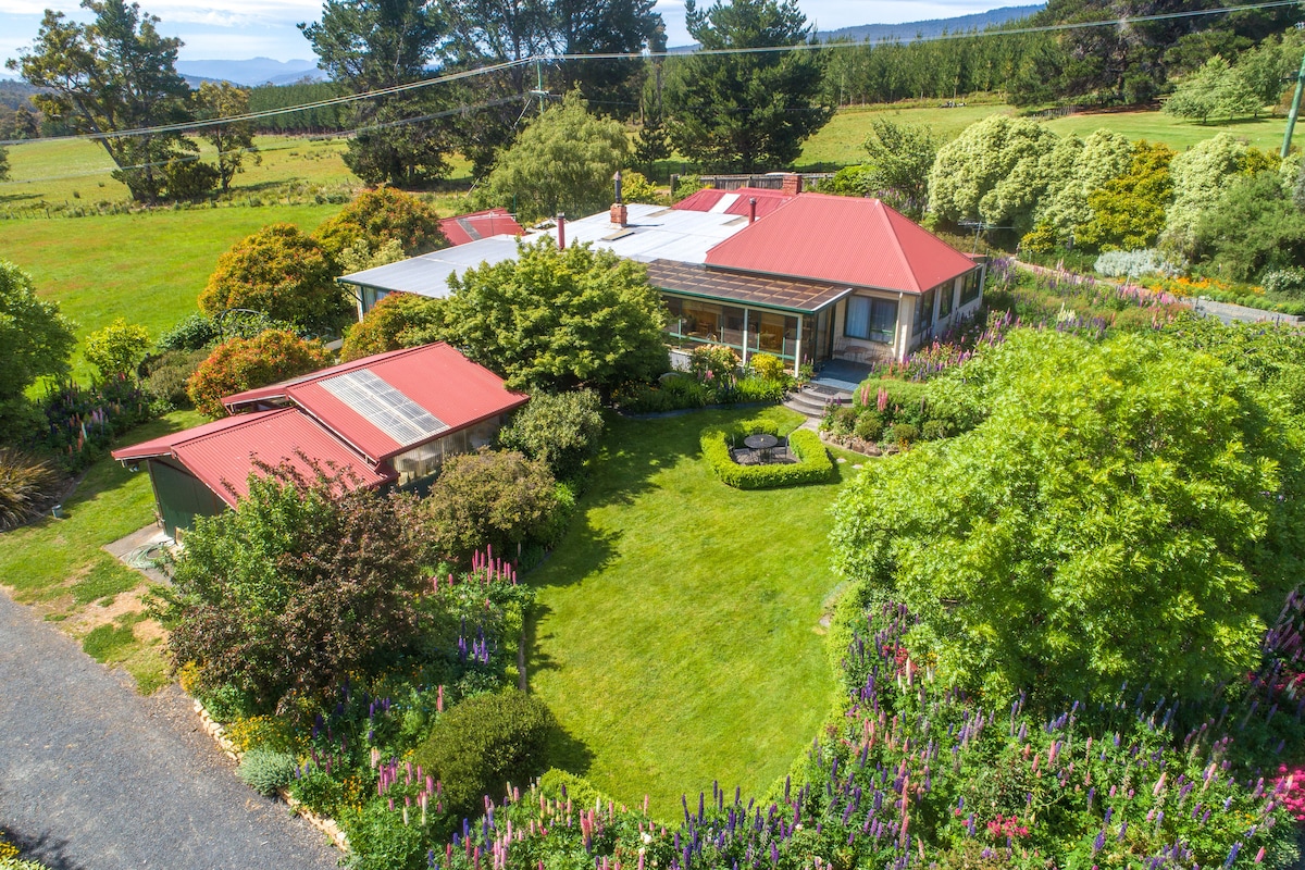 An aerial view of the property showcases a charming home with a red roof surrounded by lush gardens filled with colorful flowers. The well-maintained lawn is framed by trees and shrubs, creating a serene landscape that invites relaxation.