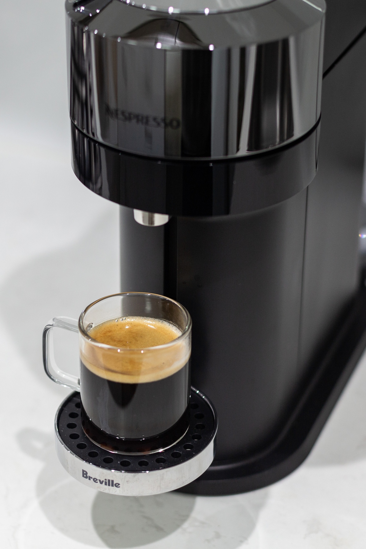A sleek black coffee machine is featured prominently, dispensing a freshly brewed espresso into a clear glass cup. The machine is positioned on a smooth, light-colored countertop, with a branded drip tray highlighting its modern design.